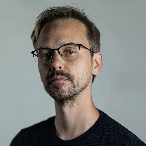 A photo of Brian, a white man with short dirty blonde hair. He has a short cropped beard and mustache, dark rimmed glasses and is wearing a black tshirt. He is looking stoicly at the camera.