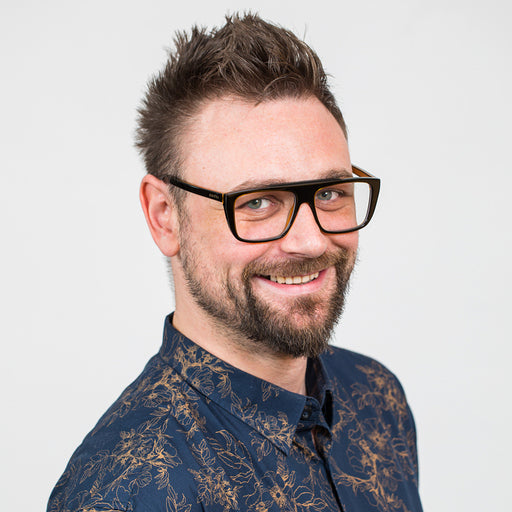 A photo of Menil a white man in his 40s in a blue collared shirt flocked with a gold pattern. He is turned 3/4 to the camera, smiling and wearing heavy rimmed glasses. He has short brown hair that is spiked upward