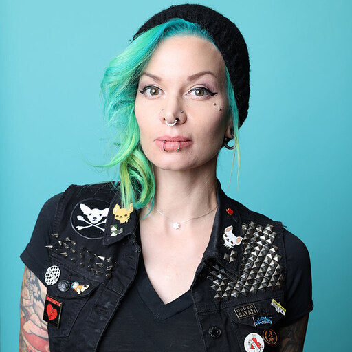 Photo of Verona, a white woman with shoulder length blue and green hair pulled over one shoulder under a black knit cap. She is wearing a black vneck t-shirt, and   a black denim vest coverd in pins and studs. She as a nose ring and two lip rings.